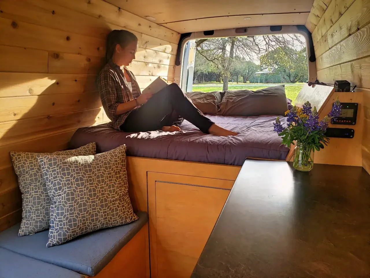 Une jeune femme lit dans un **int&eacute;rieur am&eacute;nag&eacute;** de van, entour&eacute;e de coussins et de fleurs, profitant de la lumi&egrave;re du soleil.