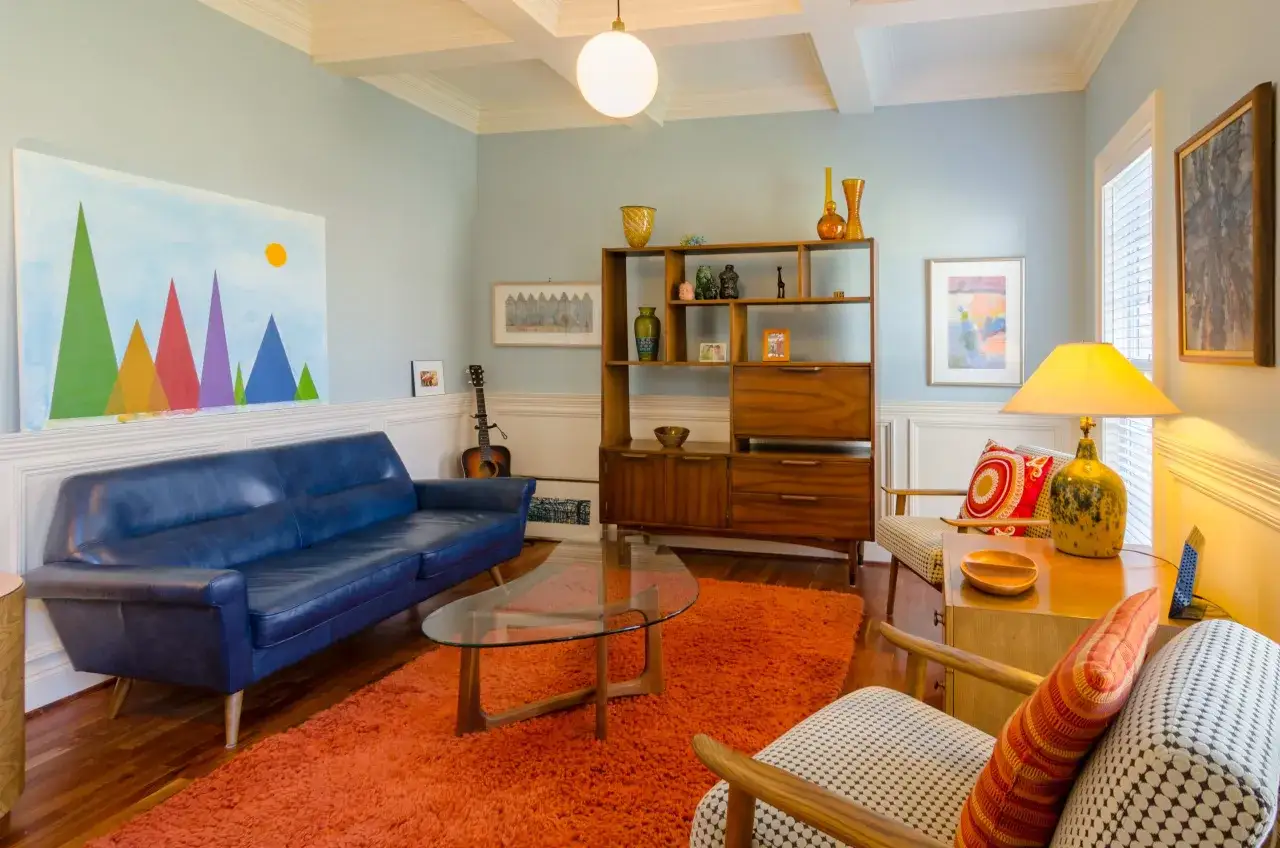Salon w stylu mid century: niebieska sofa, szklany stolik, pomarańczowy dywan, drewniana biblioteczka i kolorowe obrazy tworzą przytulną atmosferę.