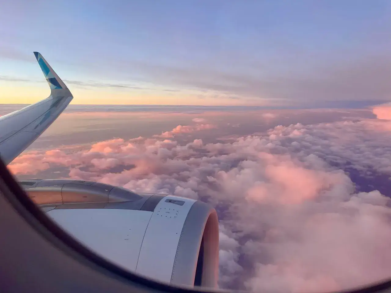 View from an airplane window at sunset. The wing and engine are visible, with pink and purple clouds stretching to the horizon.