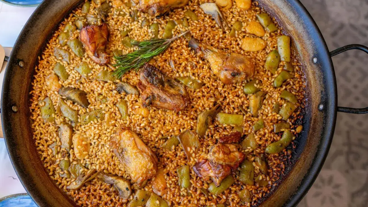 Paella valenciana recién hecha, con trozos de pollo, judías verdes y alcachofas, coronada con una ramita de romero.