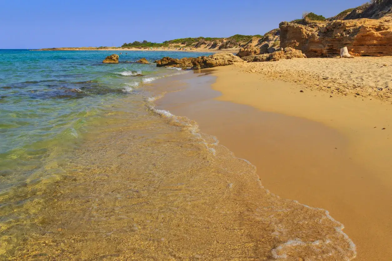 Onde cristalline lambiscono sabbia dorata su spiagge libere in Puglia, con rocce e macchia mediterranea sullo sfondo.