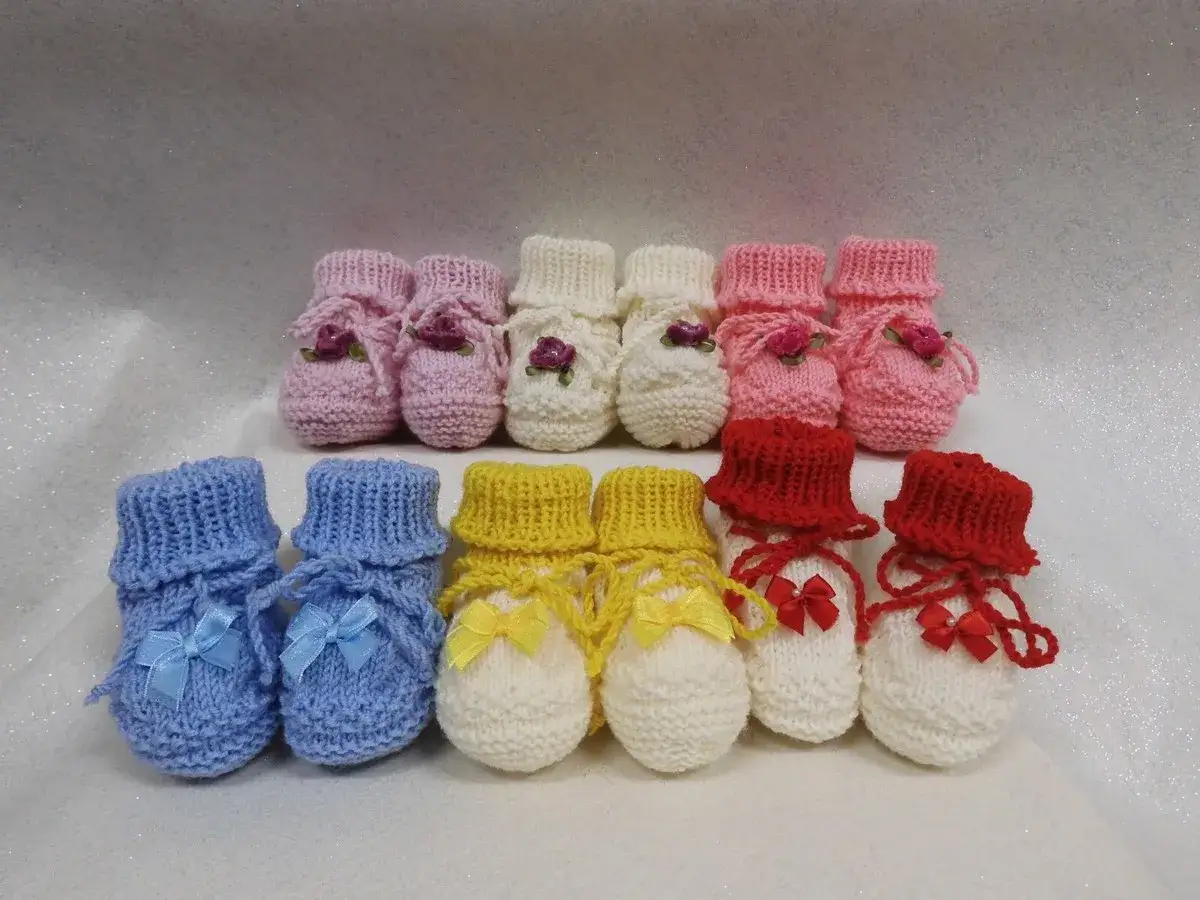 Una fila di babbucce neonato ai ferri rosa e bianche decorate con fiorellini, sotto una fila di babbucce neonato ai ferri blu, gialle e bianche con fiocchi.