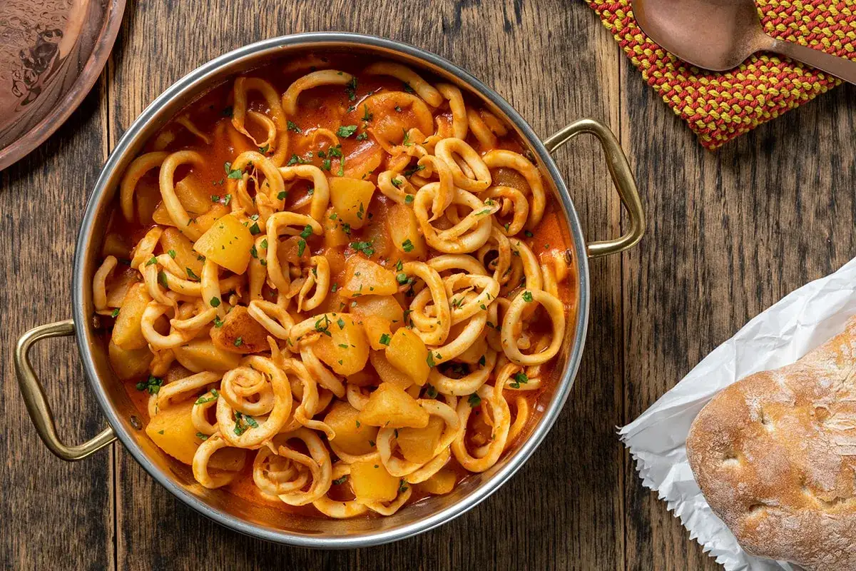 Un saporito piatto di totani e patate in umido, guarnito con prezzemolo fresco, servito in una padella di metallo su un tavolo di legno.