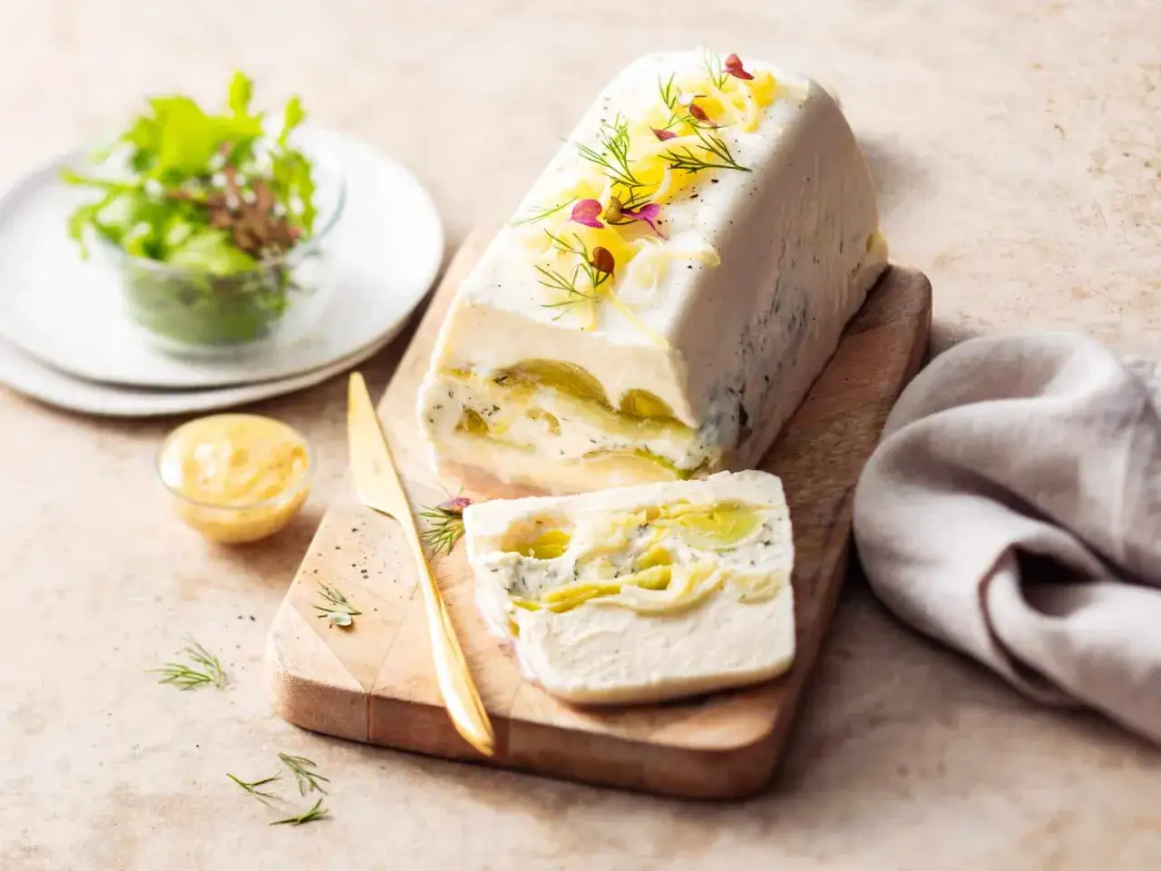 Une tranche de terrine de l&eacute;gumes, garnie d'herbes et de fleurs, repose sur une planche en bois. &Agrave; c&ocirc;t&eacute;, une petite coupelle de sauce et une salade verte.
