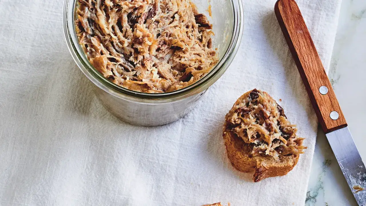 Un tarro de rillettes, una pasta de carne deshebrada, junto a una tostada untada. Un cuchillo de madera reposa al lado.