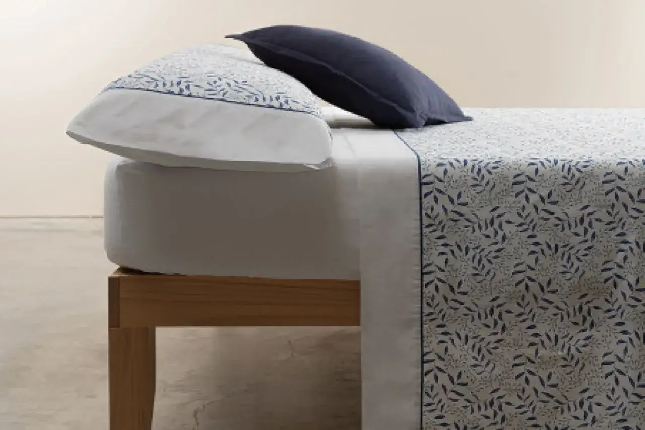 Cama con sábanas de **poliéster o microfibra** con estampado de hojas azules. Una almohada azul marino y otra con el mismo estampado.