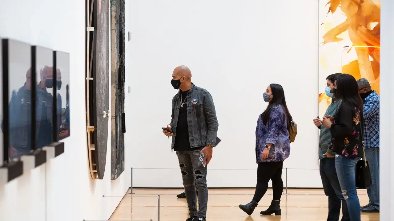 Des visiteurs masqués contemplent des œuvres d'art contemporain dans une galerie. L'un d'eux tient un téléphone, peut-être pour documenter sa visite.