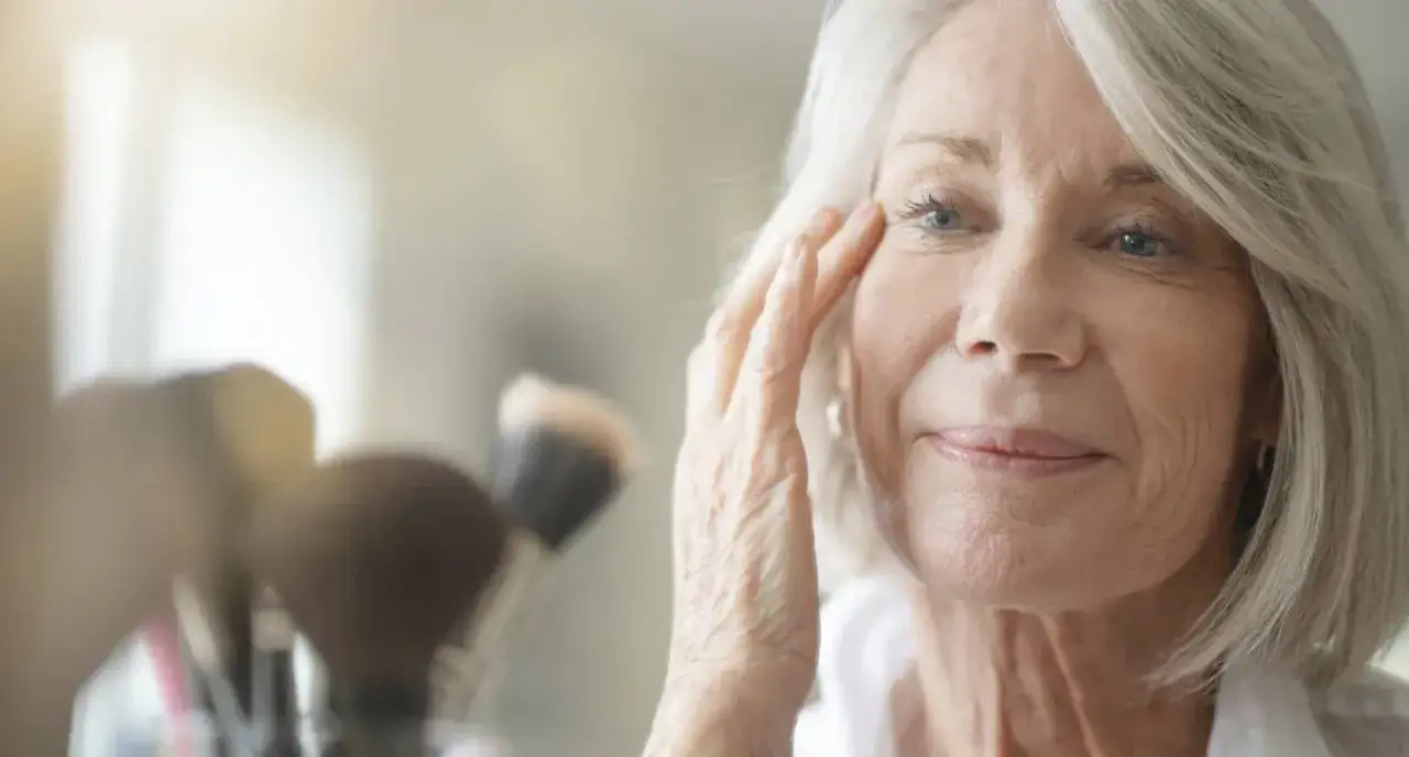 Eine Frau über 70 mit silbernem Haar und blauen Augen, die sich im Spiegel ihr Gesicht streichelt. Sie ist ein Beispiel für zeitlose Schönheit und gute Gesichtspflege.