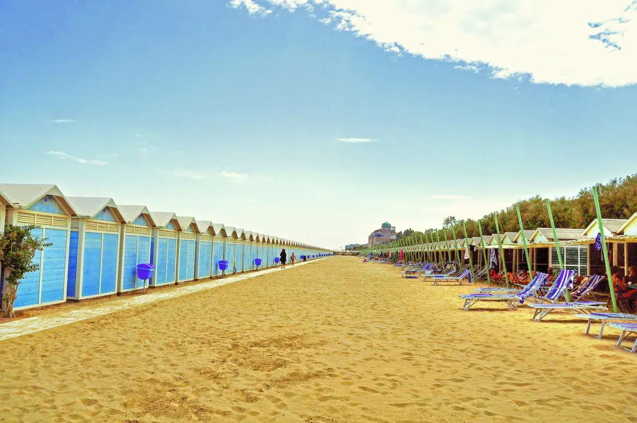 Plage de sable dor&eacute; au Lido di Venezia, avec des cabines bleues align&eacute;es d'un c&ocirc;t&eacute; et des chaises longues sous des auvents de l'autre.