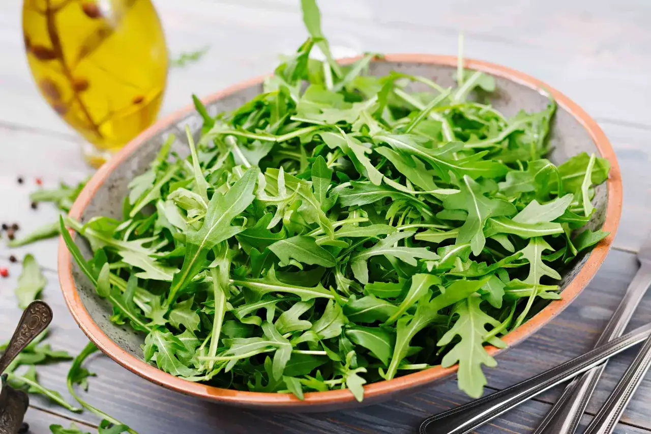 Sch&uuml;ssel voller frischem Rucola, bereit f&uuml;r ein gesundes Mittagessen. Daneben eine Flasche Oliven&ouml;l und Besteck.