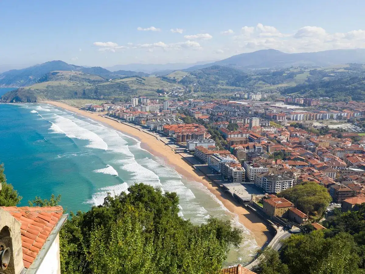 Vista panor&aacute;mica de una playa dorada con olas turquesas y una ciudad costera con tejados rojos, ideal para escapadas fin de semana Espa&ntilde;a.