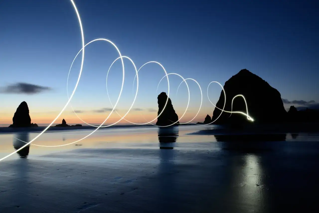 Ciel crépusculaire sur la plage, avec des rochers découpés en silhouette. Des traînées lumineuses, créées par des photos pose longue, dessinent des spirales sur l'eau calme.