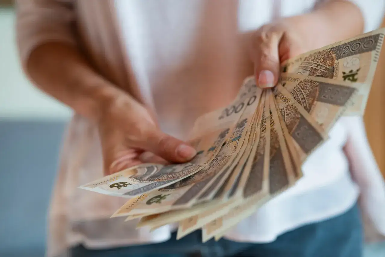 Dłonie trzymają wachlarz banknotów 200 zł. To symbol nagrody jubileuszowej za 20 lat pracy, docenienie długoletniego stażu.