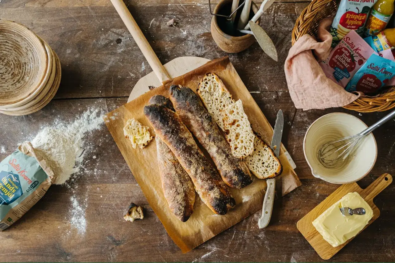 Nybakade surdegsbaguetter p&aring; bakpl&aring;tspapper, redo f&ouml;r ett surdegslimpa recept. Mj&ouml;l, sm&ouml;r och ingredienser f&ouml;r bakning ligger utspridda.