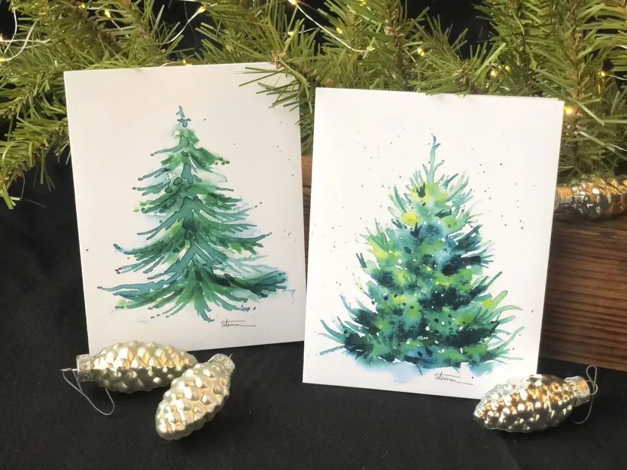 Dos tarjetas navide&ntilde;as manualidades con acuarelas de &aacute;rboles de Navidad, adornos plateados en forma de pi&ntilde;a y ramas de abeto con luces.