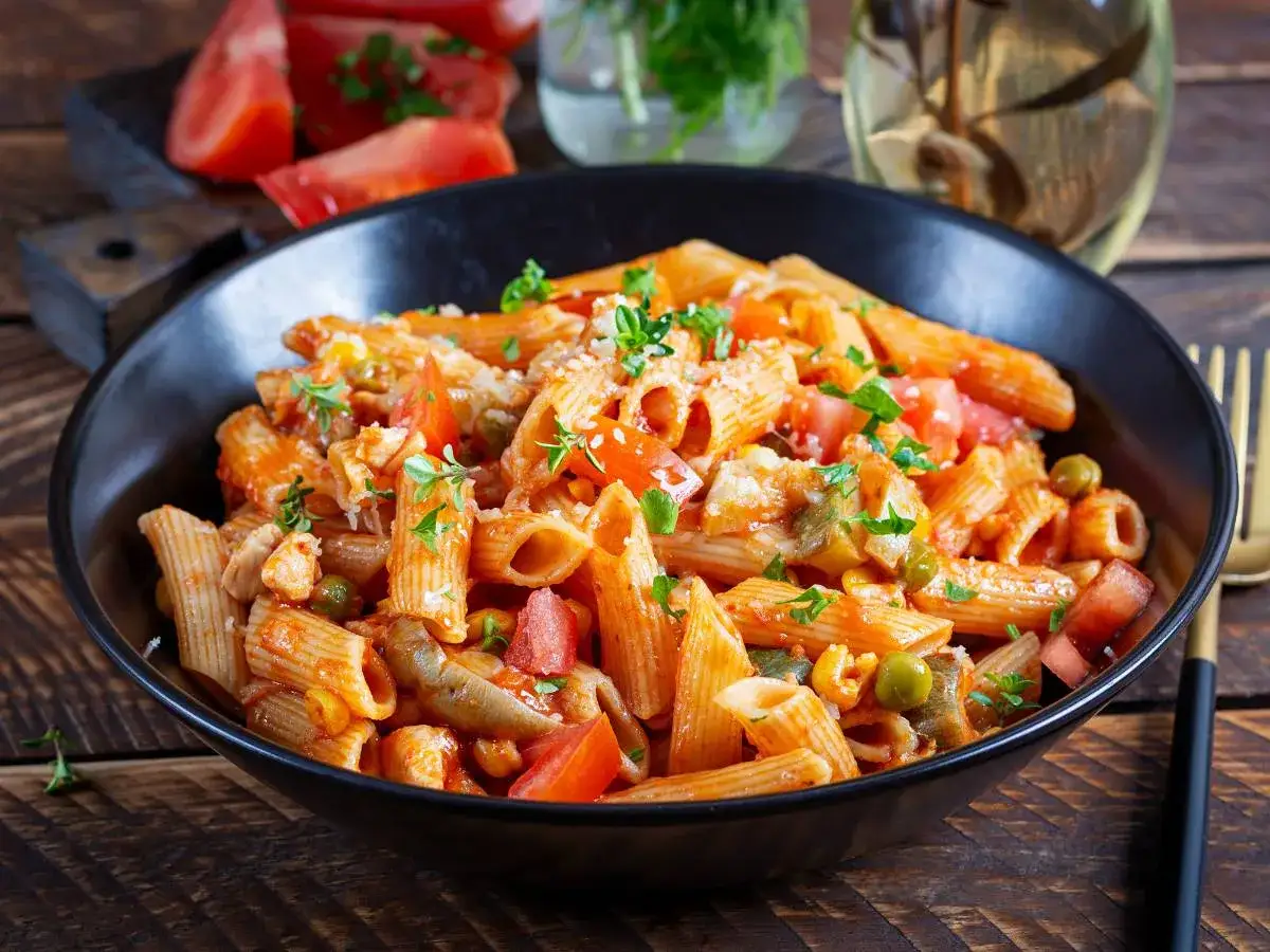 Penne w pikantnym sosie pomidorowym z kawałkami kurczaka, groszkiem i pomidorami. Danie posypane natką pietruszki, wygląda jak pyszne spaghetti arrabiata.