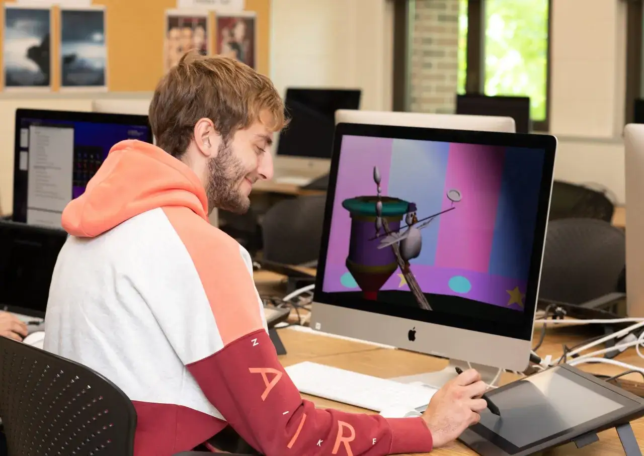 Ein junger Mann mit Bart arbeitet an seinem Computer an einer Animation. Er nutzt ein Grafiktablett, um die Figur eines Vogels zu gestalten, der auf einem Stab balanciert.