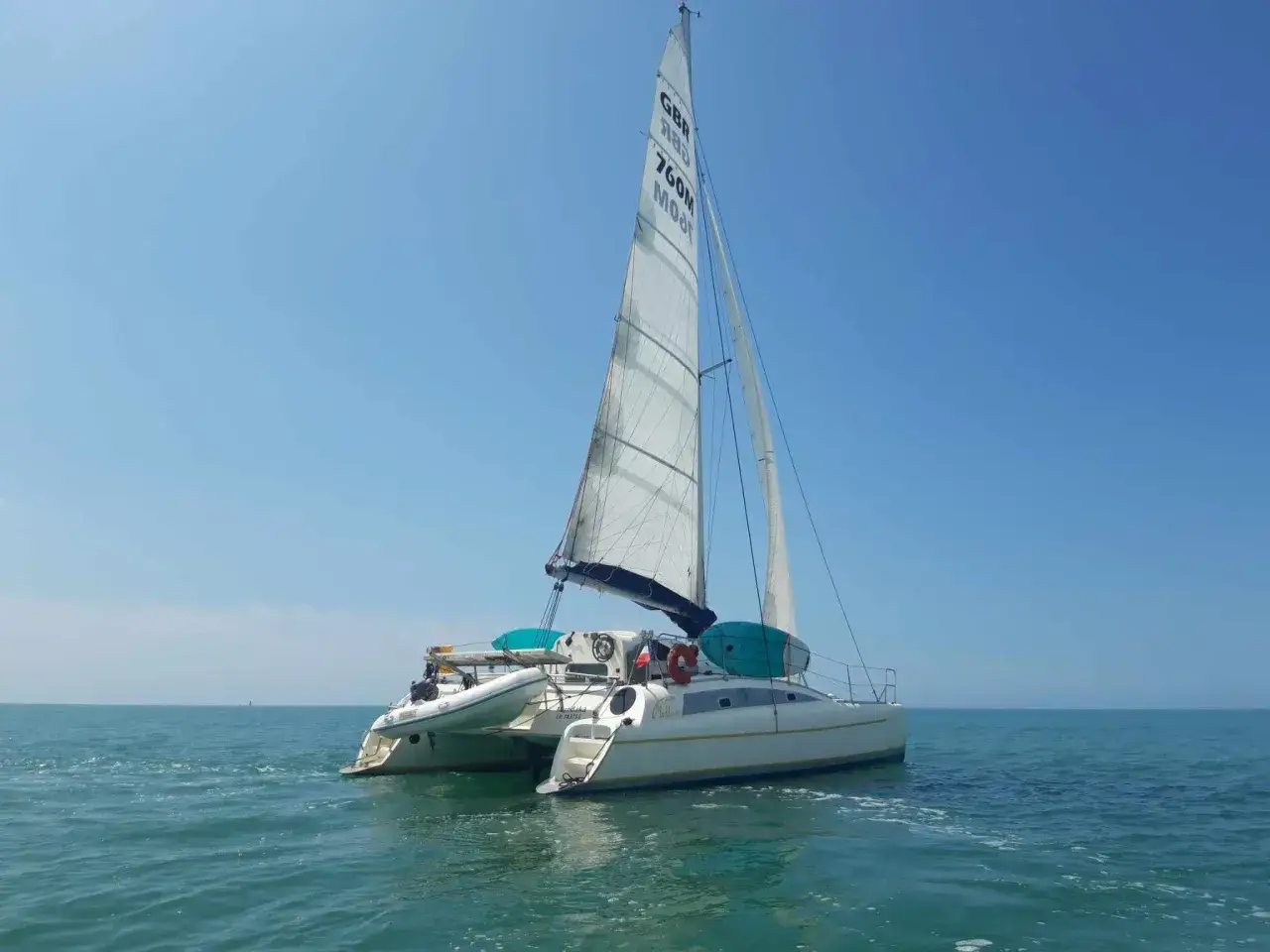 Un petit catamaran habitable navigue sur une mer calme sous un ciel bleu clair.