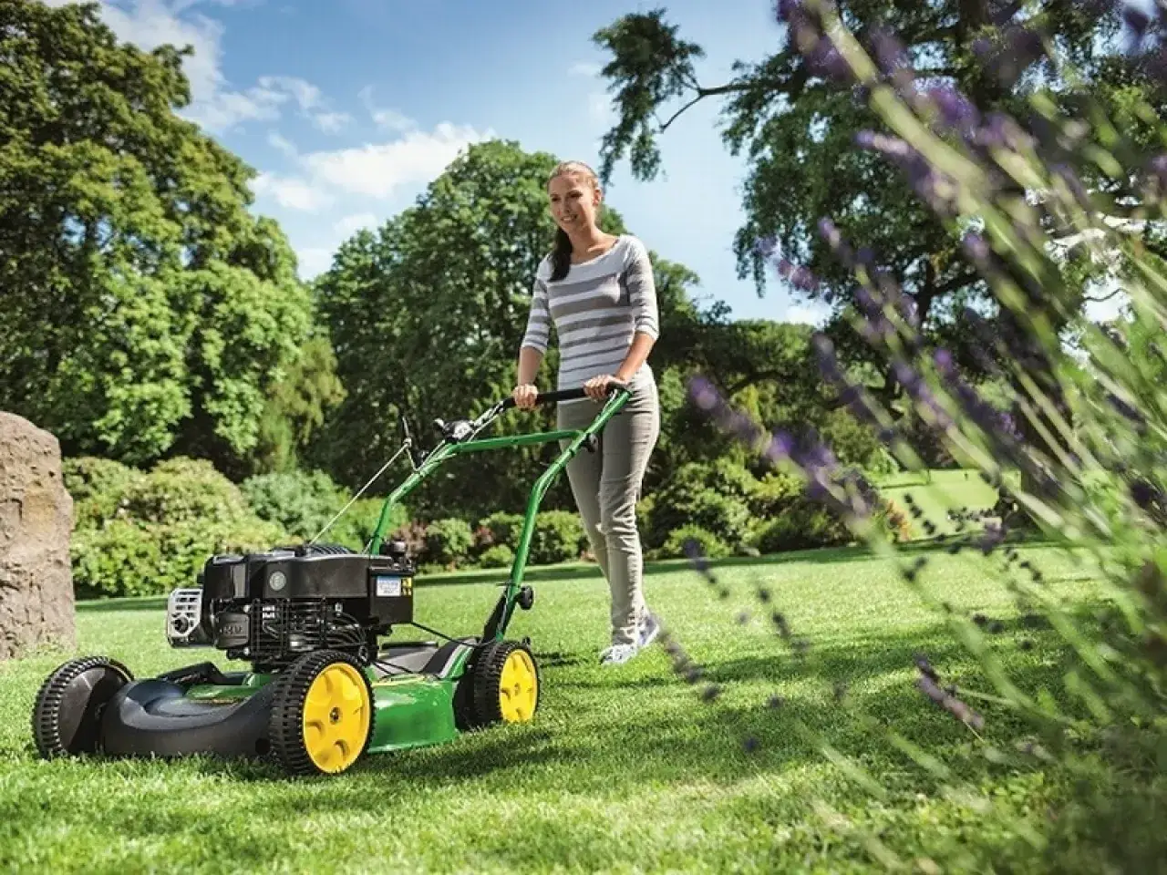 Uśmiechnięta kobieta pcha zielony mulczer do trawy po zadbanym trawniku, otoczonym bujną zielenią i kwitnącą lawendą.
