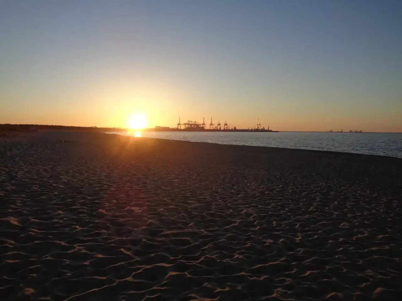 Piękny zachód słońca nad Bałtykiem w Gdańsku. Słońce nisko nad horyzontem, złocące plażę i morze. W oddali widać portowe dźwigi.