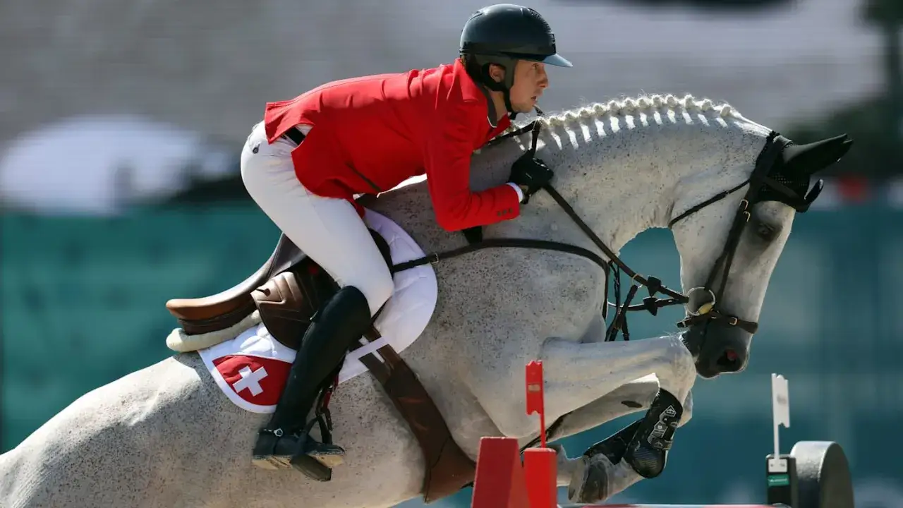 Cavalier en tenue rouge et blanc, sur un cheval gris moucheté, en plein saut. Une image qui illustre la **FEI definition** de l'équitation de haut niveau.