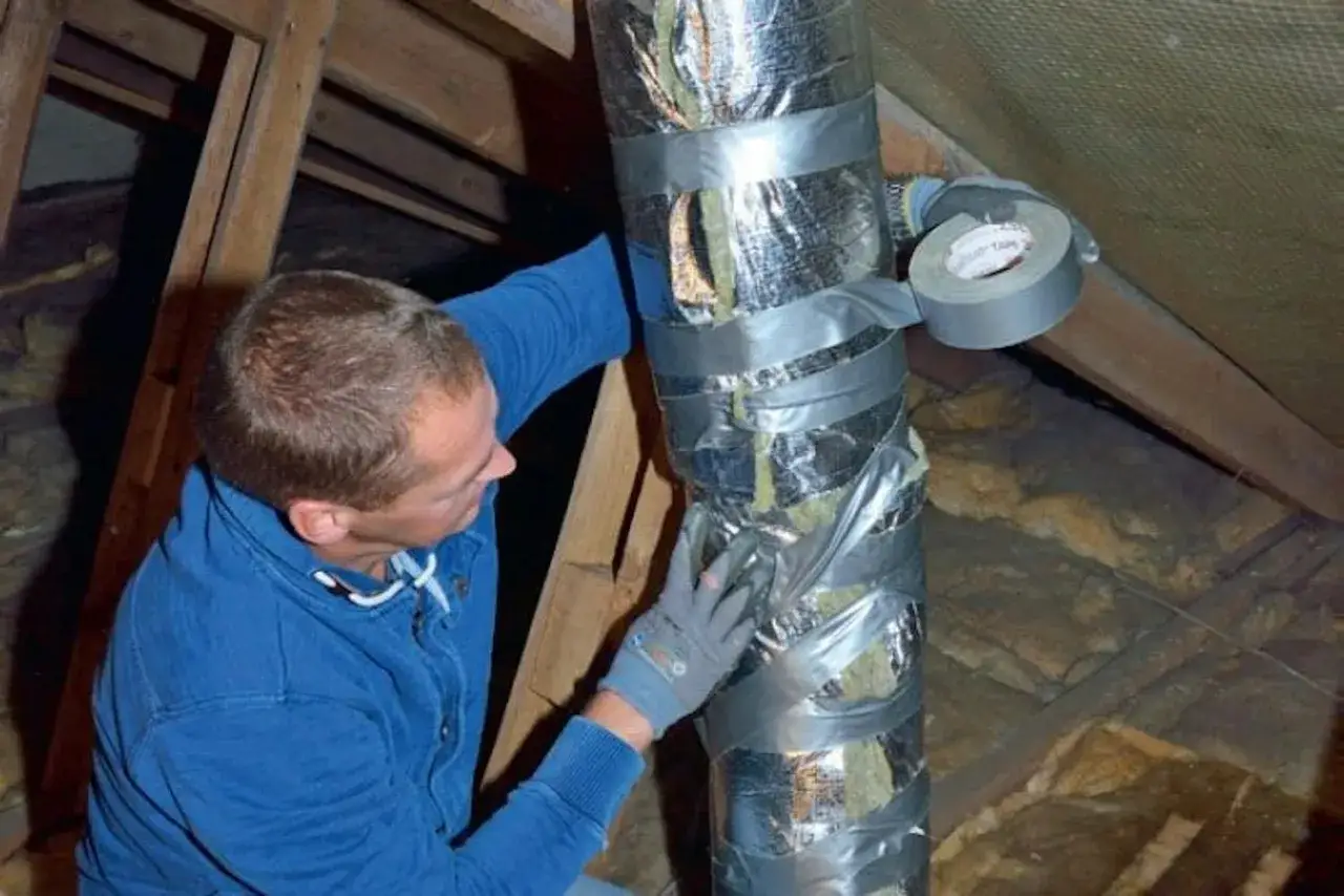 Man i blå tröja och handskar tejpar isolerad ventilationskanal i ett vindsutrymme. Han verkar byta syll.