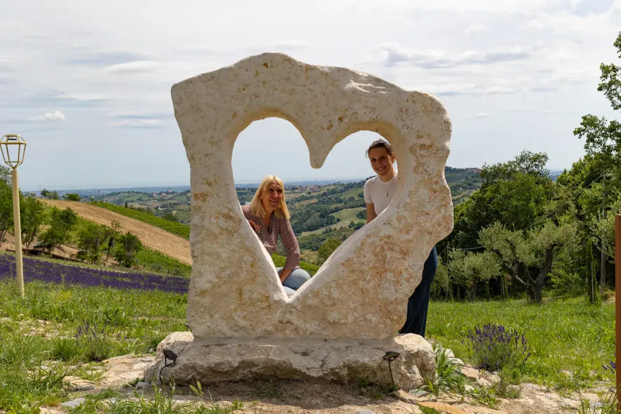 Due donne sorridono dietro un cuore di pietra, con il lavandeto di Monteleone e dolci colline sullo sfondo.