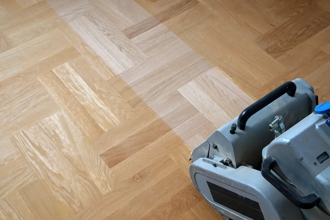 Maskin slipar parkettgolv i fiskbensm&ouml;nster. En del av golvet &auml;r ljusare d&auml;r slipningen p&aring;g&aring;r.