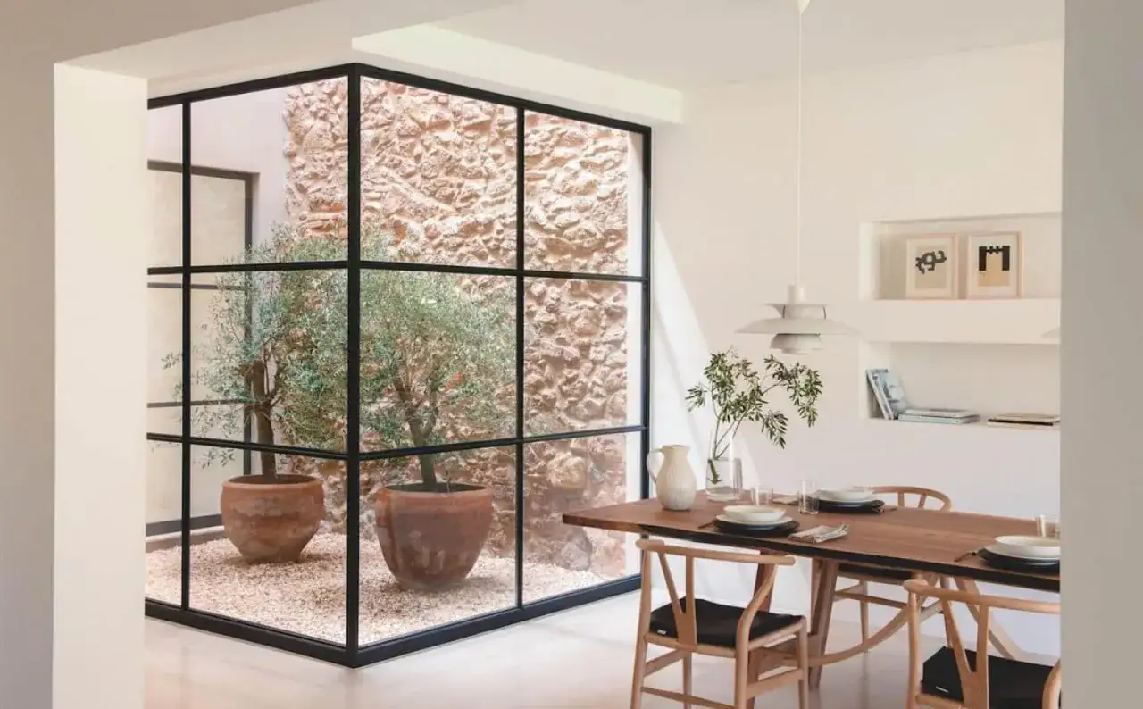 Un comedor moderno con una mesa de madera y sillas de dise&ntilde;o, junto a un patio de luces con olivos en macetas y paredes de piedra.