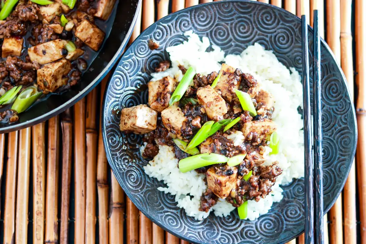 En sk&aring;l med &aring;ngande ris toppad med mapo tofu och garnerad med salladsl&ouml;k. En panna med mer mapo tofu syns till v&auml;nster.