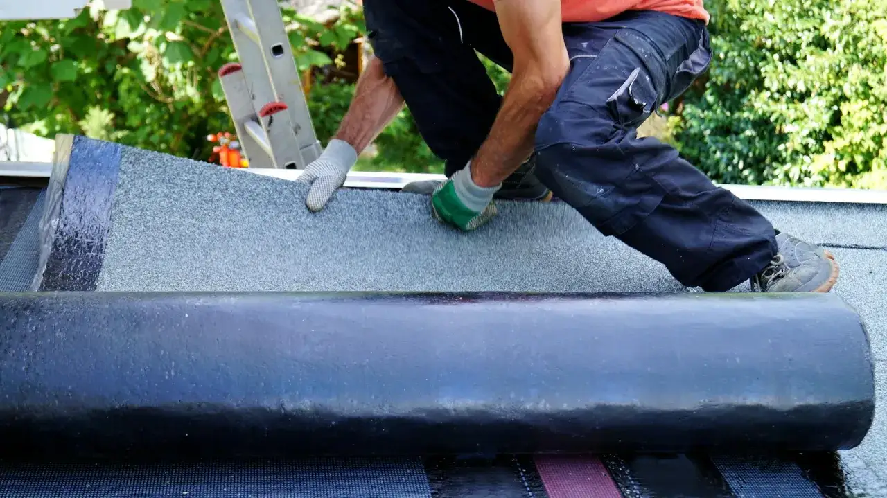 Handwerker mit Handschuhen legt Dachpappe an. Die Rolle mit der neuen Dachpappe liegt bereit, um das Dach sicher zu machen.