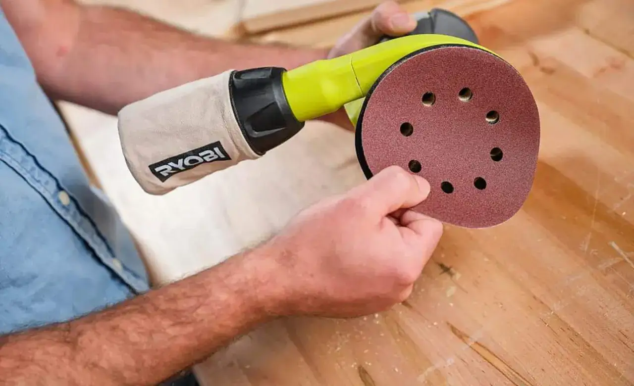 Un homme utilise un outil pour poncer du bois. Le disque de ponçage est rouge et le sac de récupération de poussière porte le logo Ryobi.