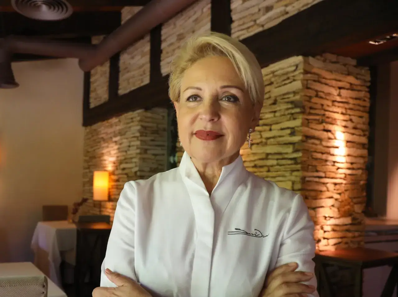 Susi Díaz, chef con su distintiva firma bordada en su uniforme blanco, posa con confianza en un restaurante rústico.