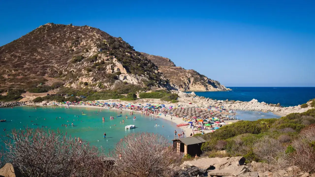 Una baia affollata a Punta Molentis Beach, con acque turchesi, sabbia bianca, ombrelloni colorati e persone che nuotano e prendono il sole.