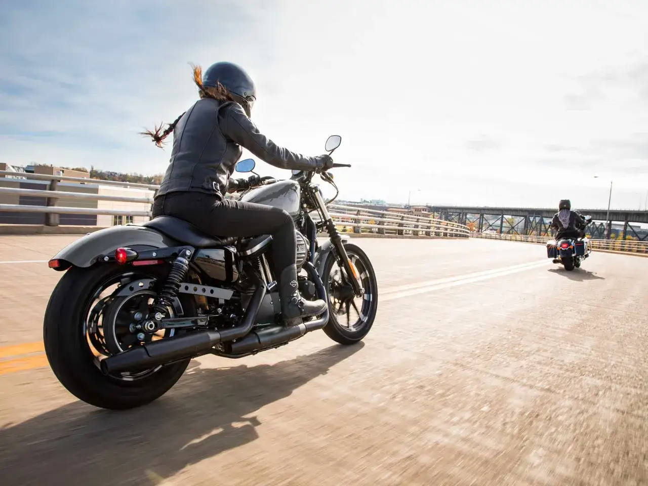 Kobieta w skórzanej kurtce i kasku jedzie na stylowym motocyklu dla kobiety. Wiatr rozwiewa jej włosy, a słońce oświetla drogę.