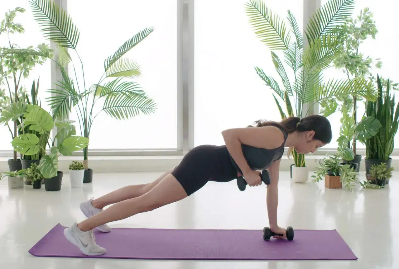 Femme en position de planche, effectuant un exercice dos avec halt&egrave;res. Entra&icirc;nement &agrave; domicile entour&eacute;e de plantes.