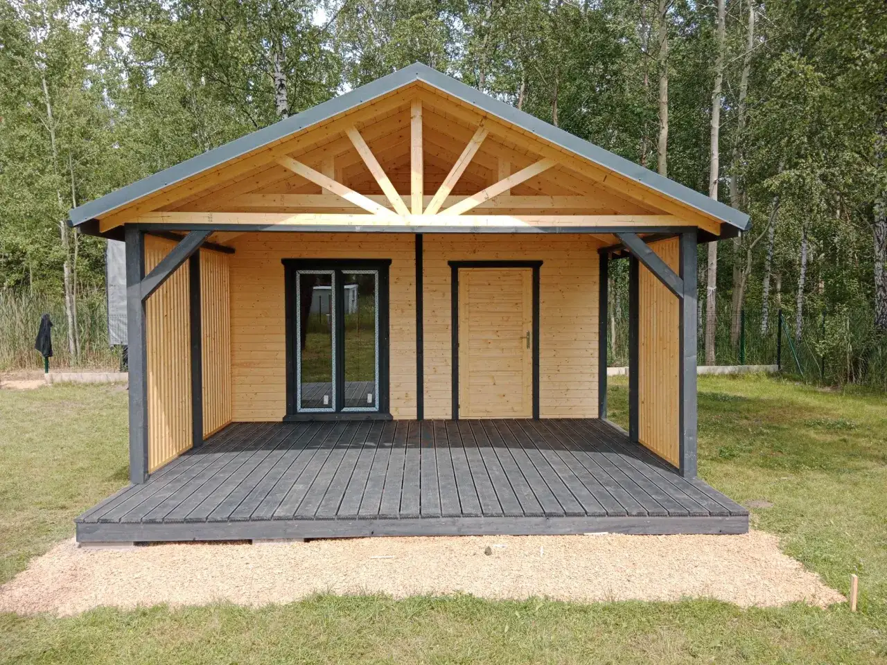 Gotowy domek letniskowy z drewnianym tarasem i przeszklonymi drzwiami, otoczony zielenią.