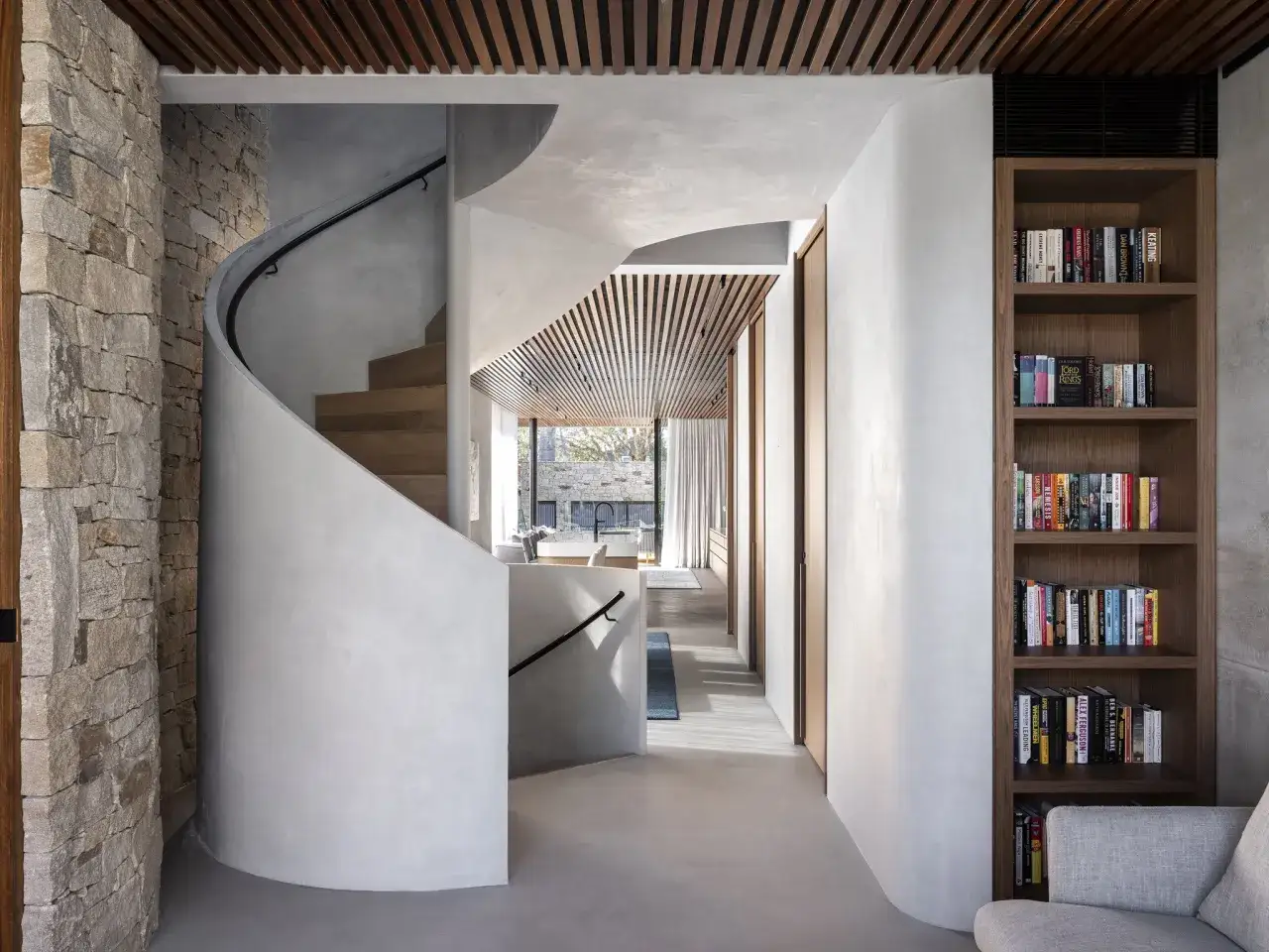 Escalera de caracol con revestimiento de microcemento, piedra y madera. Estanter&iacute;a llena de libros.
