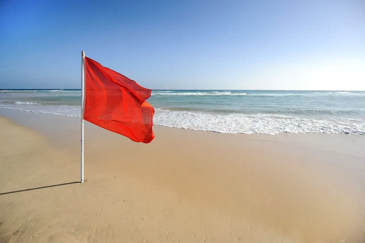 Czerwona flaga powiewa na wietrze na pustej plaży. Morze jest spokojne, ale niebo jest czyste.