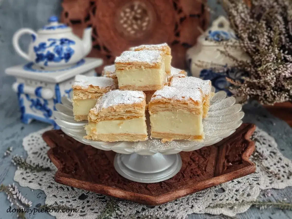 Kremowy tort napoleon, posypany cukrem pudrem, prezentuje się apetycznie na paterze. W tle ozdobny dzbanek i wrzosy.