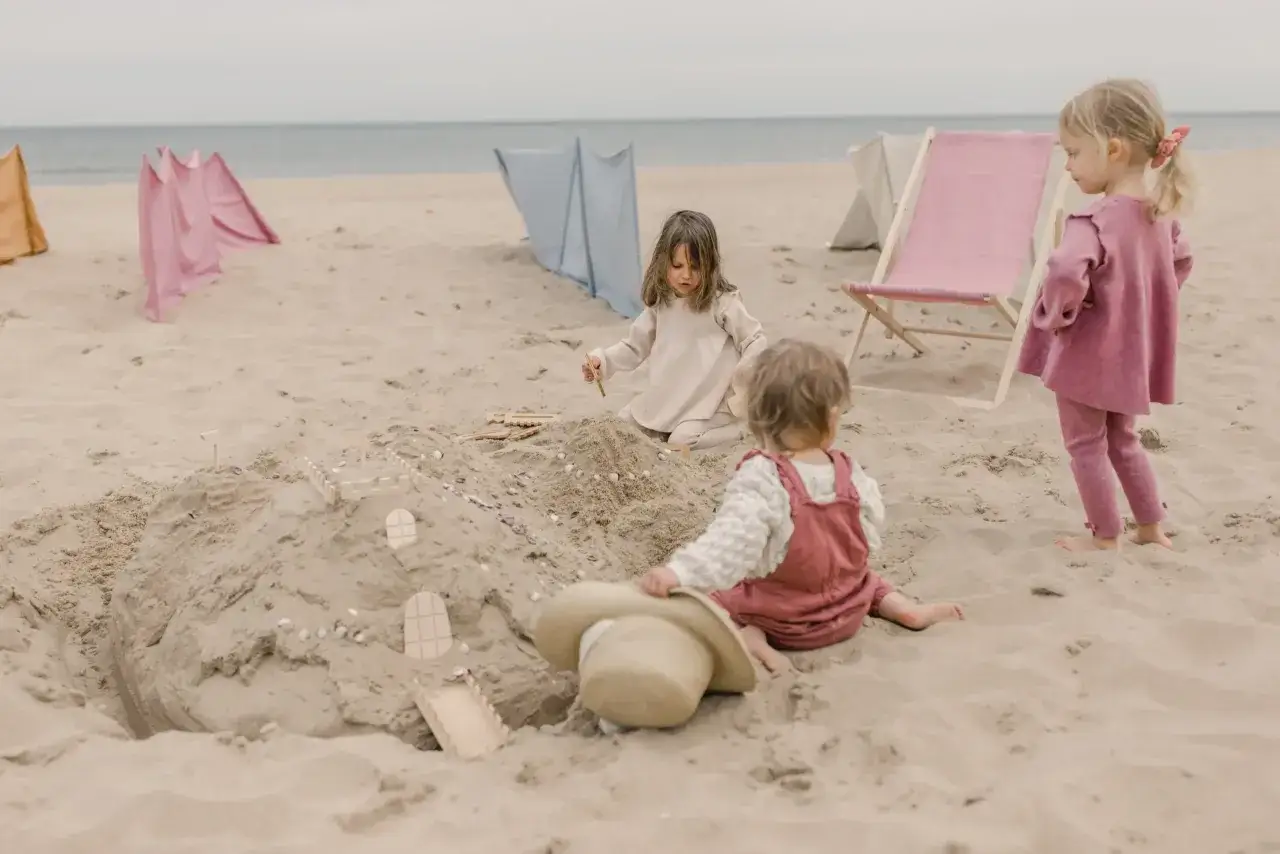 Dzieci budują zamek z piasku, ciesząc się beztroskimi zabawami na plaży. W tle widać leżaki i parawany.
