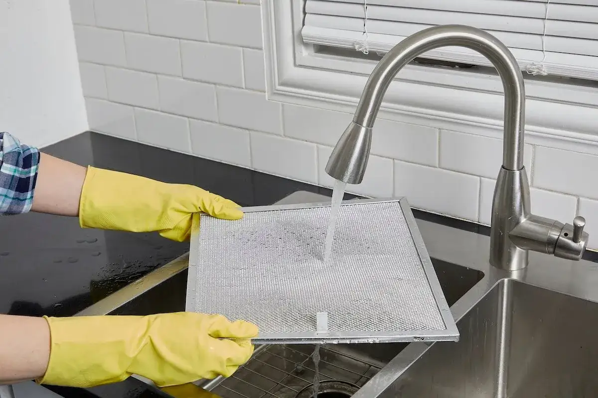 Hands in yellow gloves rinse a greasy vent hood filter under a faucet. Cleaning it regularly prevents fire hazards and ensures better air quality.
