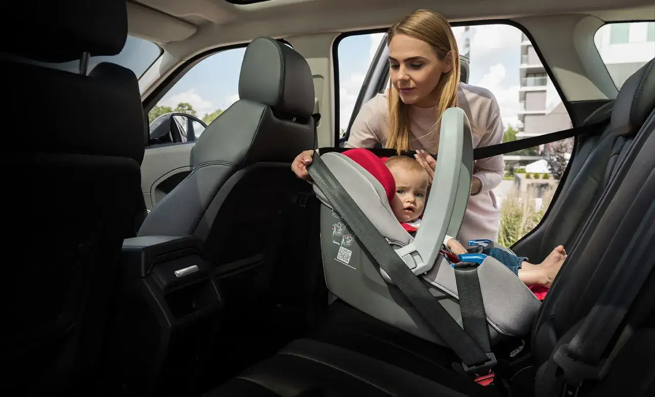 Mama pokazuje, jak zapiąć nosidełko pasami w samochodzie. Dziecko siedzi bezpiecznie w foteliku.