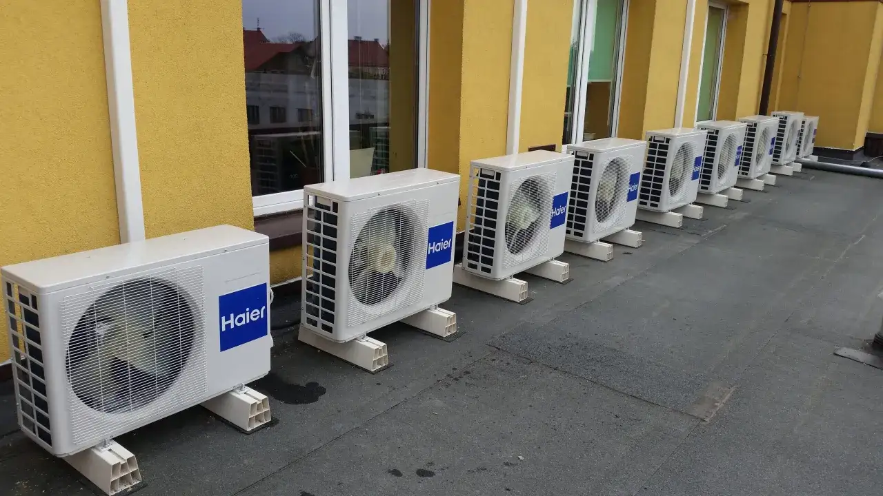 Rząd białych jednostek klimatyzacyjnych Haier na dachu, obok żółtej ściany budynku.