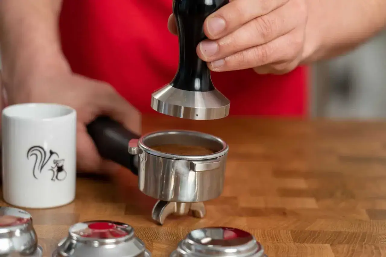 Barista dr&uuml;ckt Kaffeepulver im Siebtr&auml;ger richtig tampen, um perfekten Espresso zuzubereiten. Eine kleine Tasse mit Eichh&ouml;rnchen-Motiv steht daneben.
