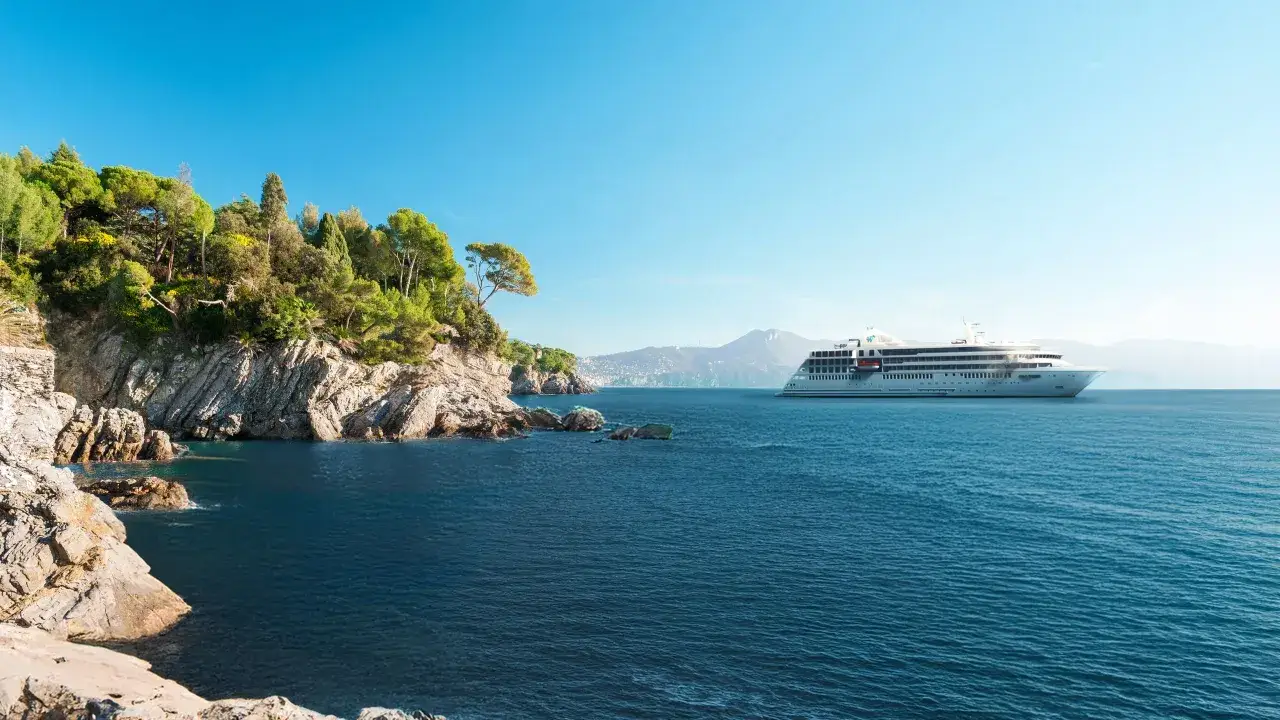 Una nave da crociera solca acque blu vicino a una costa rocciosa e verdeggiante, promettendo le migliori crociere.