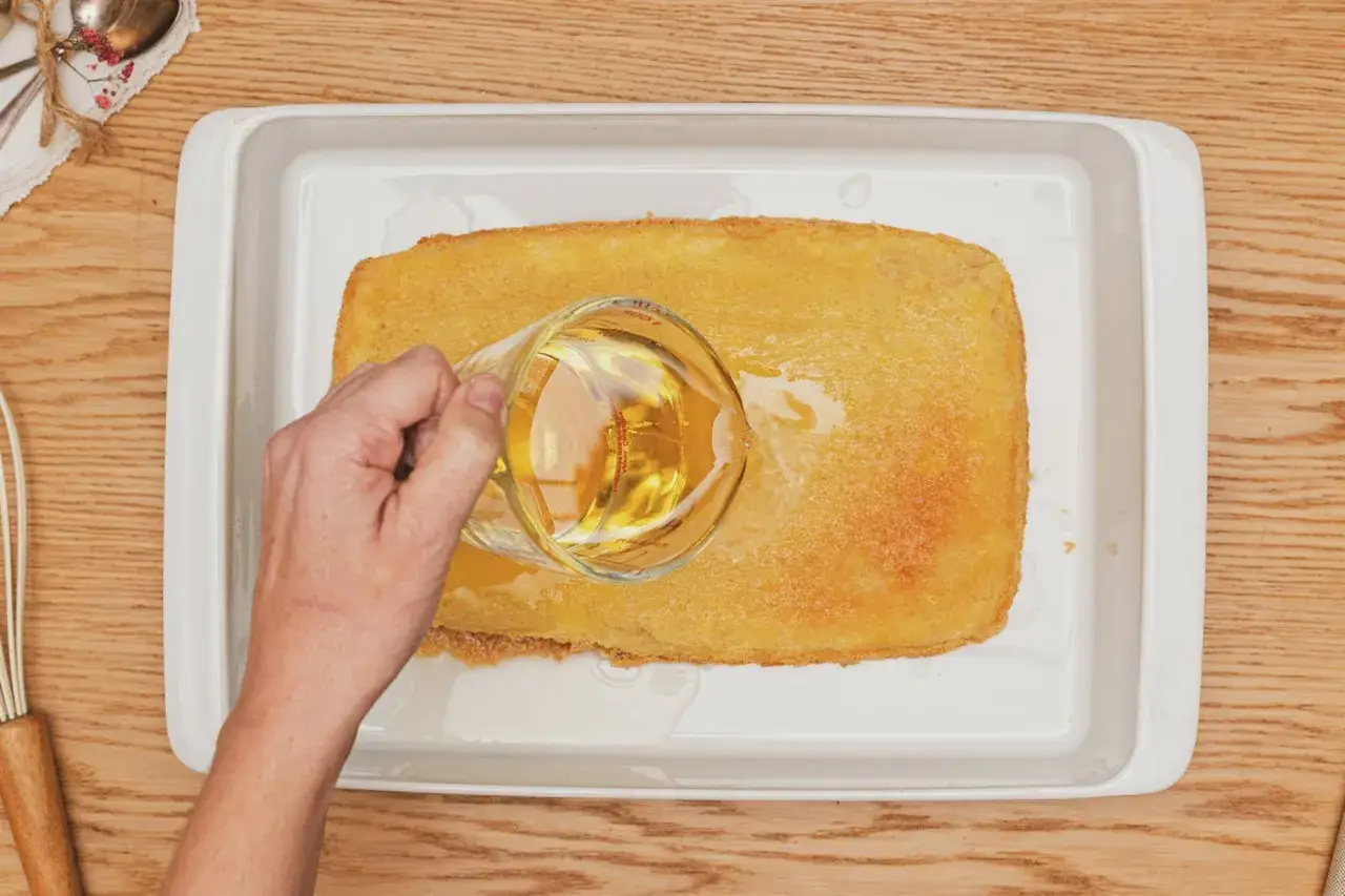 Mano vierte líquido dorado sobre bizcocho borracho en bandeja blanca.