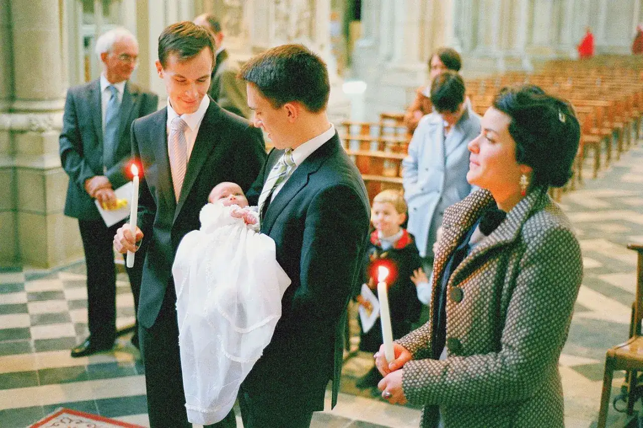Un bébé en tenue blanche est tenu par un homme lors d'un baptême religieux. Une bougie allumée est tenue par un autre homme.