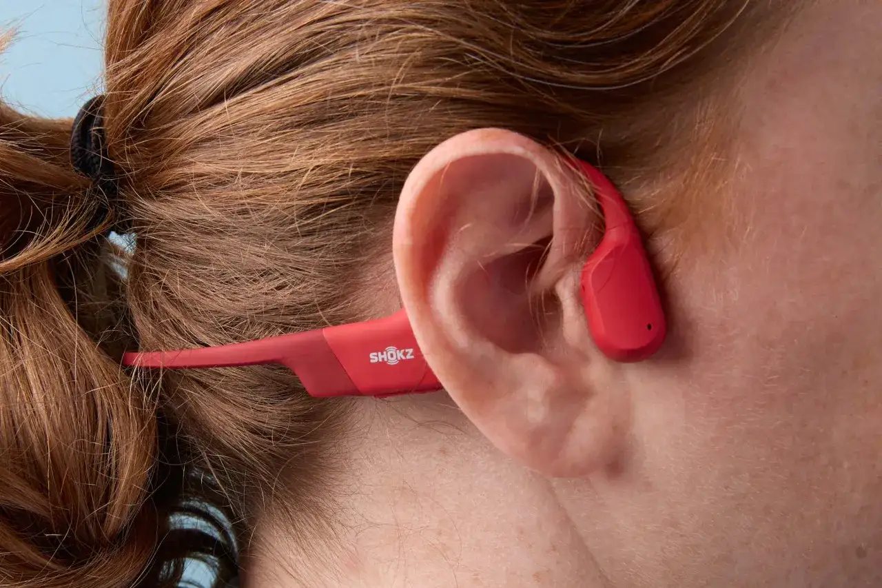 Un casque à conduction osseuse rouge Shokz, discret et léger, est porté par une personne aux cheveux châtains.