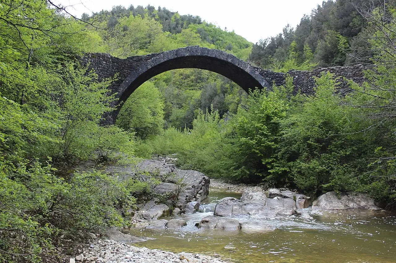 Antico ponte della Pia con un fiume che scorre tra rocce e vegetazione lussureggiante.